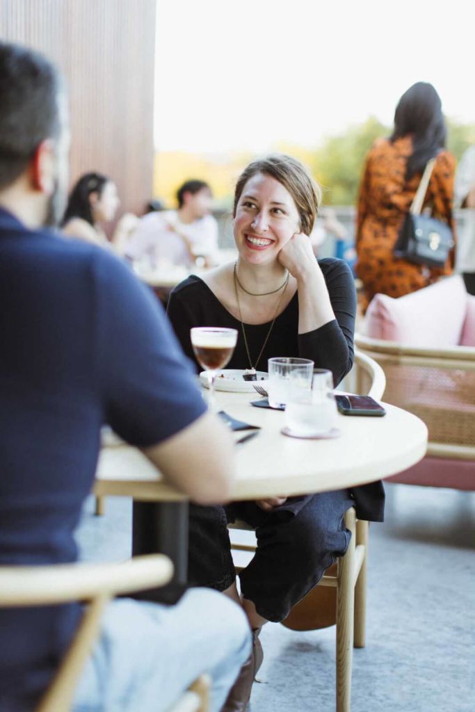 Woman smiling while sitting at a table outdoors in a patio.