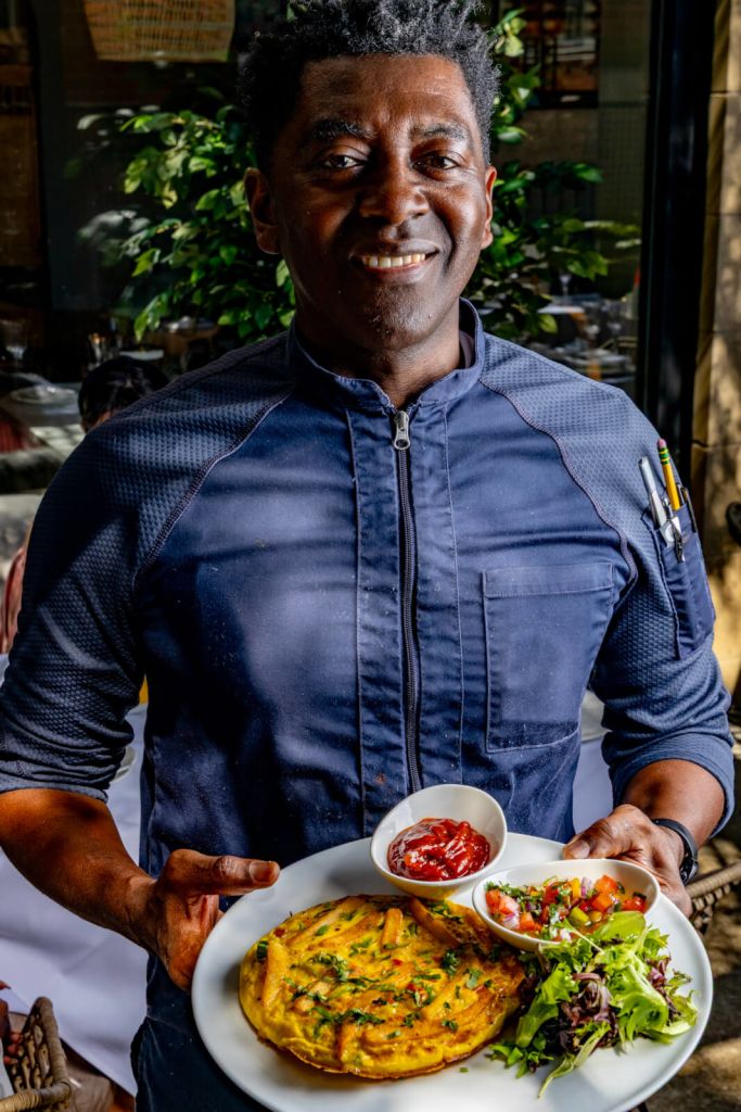 A smiling man in a blue chef's jacket holds a plate with a colorful meal, including a yellow omelette, fresh salad, and condiment bowls in Atlanta, Georgia.