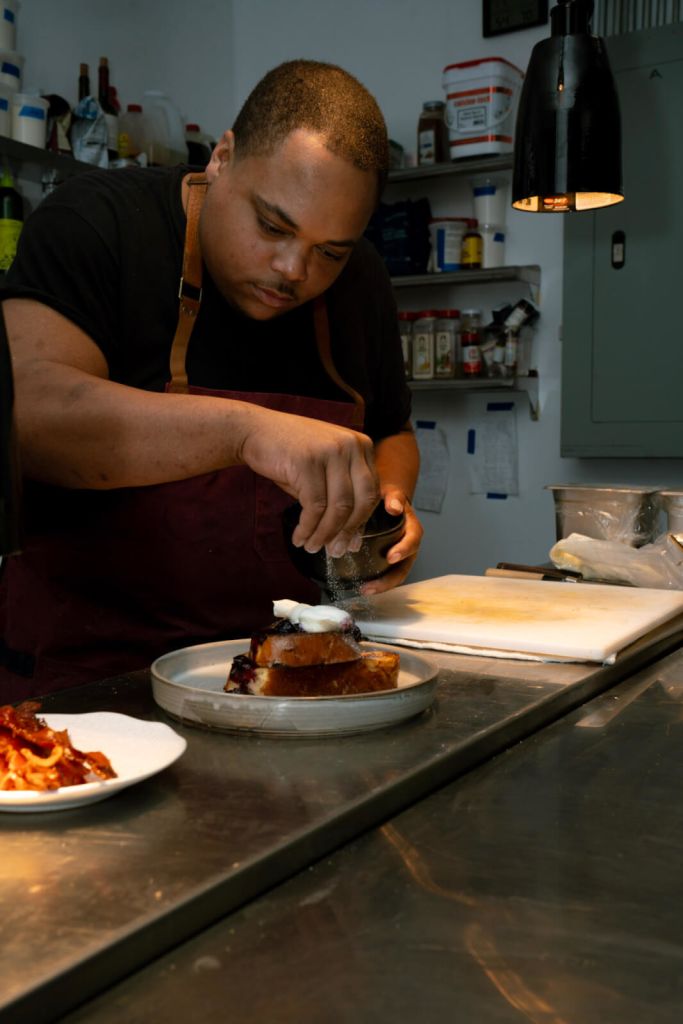 A chef in a kitchen, wearing an apron, carefully sprinkles sugar on a dish of French toast topped with whipped cream and berries in Atlanta, Georgia.