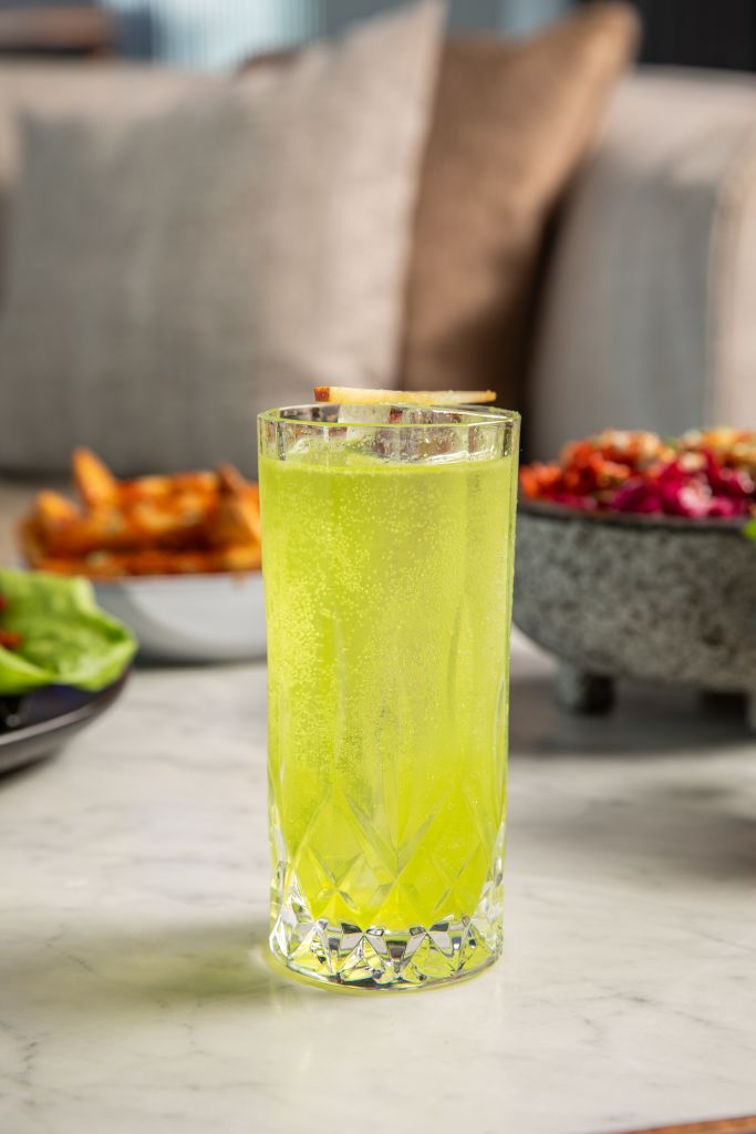 A refreshing green cocktail garnished with a lemon twist sits in a tall, crystal glass on a marble table, accompanied by a bowl of vibrant salad and crispy fries in the background in Atlanta, Georgia.