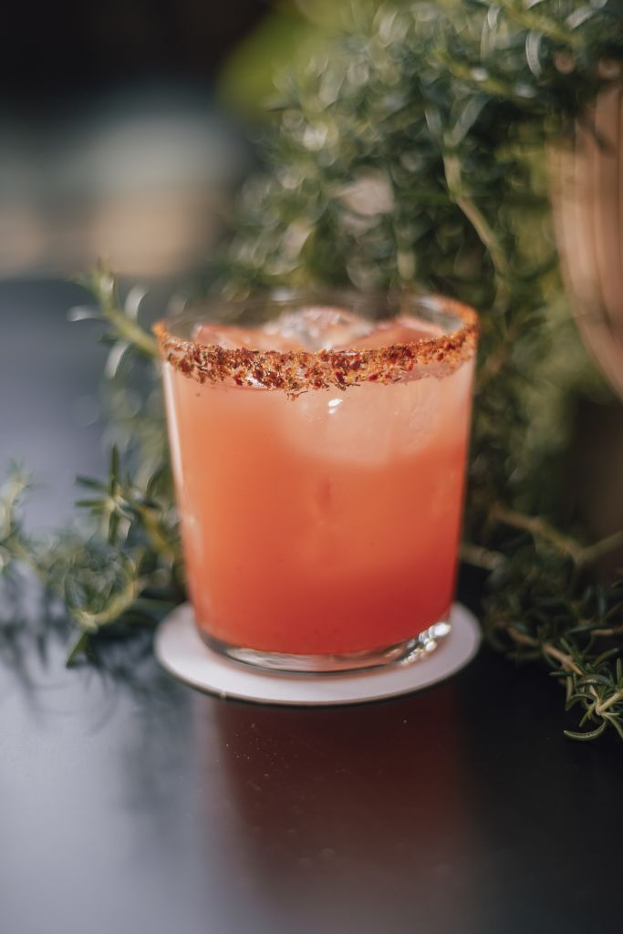 Must-Try Summer Cocktails in Atlanta 2 A vibrant orange cocktail with a rim coated in red chili powder sits on a white coaster, surrounded by fresh green herbs in Atlanta, Georgia.