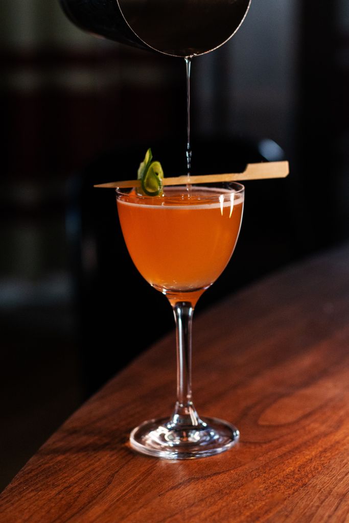 A vibrant orange cocktail is being carefully poured into a classic coupe glass, garnished with a lime wedge and a skewer in Atlanta, Georgia.