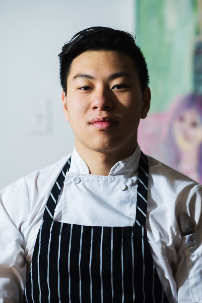 A young Asian chef with a serious expression wears a white chef's jacket and black apron in Atlanta, Georgia.