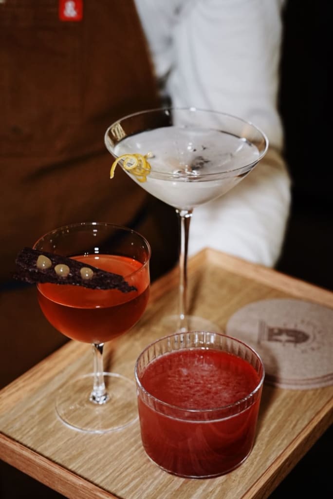 Three distinct cocktails are artfully arranged on a wooden tray: a vibrant red drink garnished with dark chocolate and golden beads, a creamy white martini with a lemon twist, and a frothy red beverage in a short glass in Atlanta, Georgia.