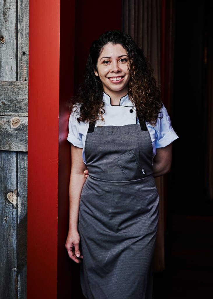 La historia detrás de Bar ANA en Eastside, una carta de amor a la cultura, la artesanía y la comunidad 1 A smiling chef in a white shirt and gray apron stands against a red wall in Atlanta, Georgia.