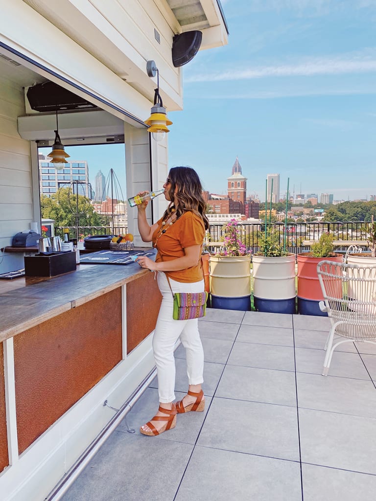 A woman in a colorful outfit enjoys a drink at an outdoor rooftop bar with a city view in Atlanta, Georgia.