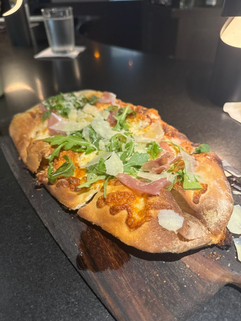 A rectangular pizza with arugula, prosciutto, and shaved Parmesan on a wooden board in Atlanta, Georgia.