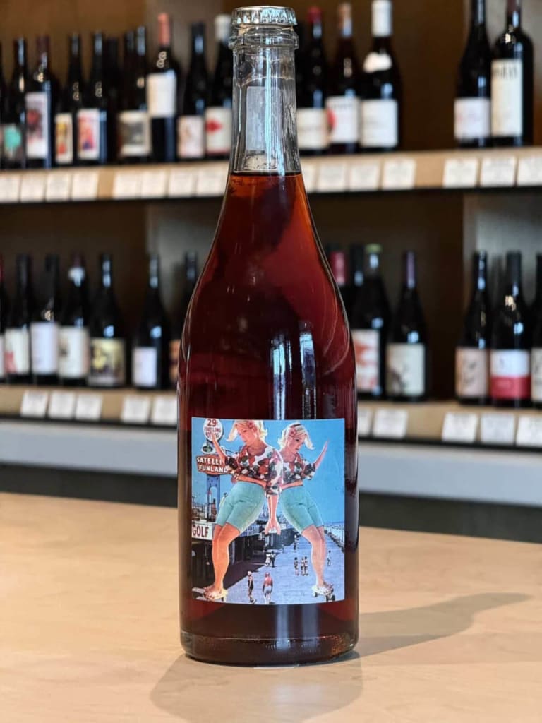A bottle of red wine with a vibrant label featuring two women in colorful outfits stands out on a wooden counter, with shelves of various wine bottles in the background in Atlanta, Georgia.