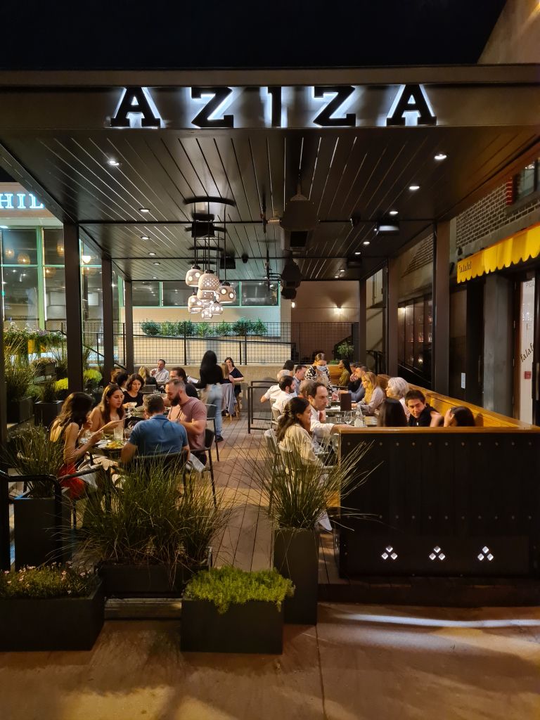 Patio under a sign that reads Aziza. People sitting at tables.
