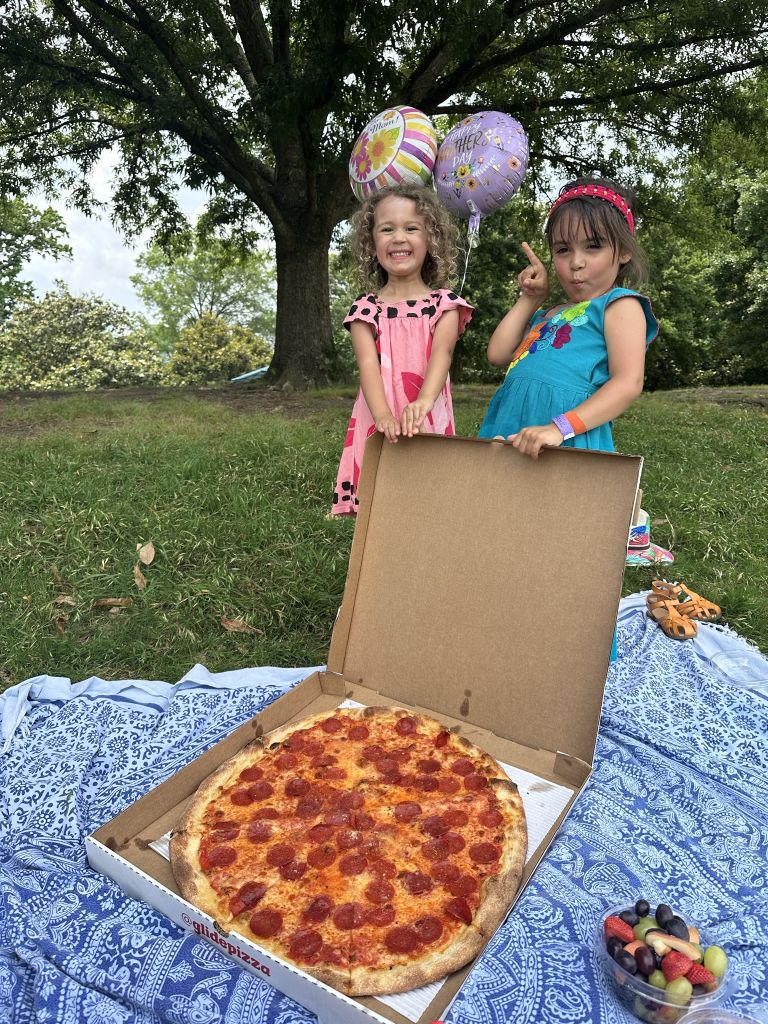 Two young girls in colorful dresses hold balloons above a large pepperoni pizza on a picnic blanket in Atlanta, Georgia.
