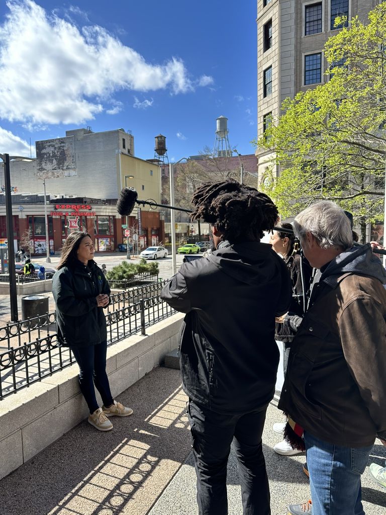 A group of people, including a woman in a black jacket and a man with dreadlocks, are gathered on a sidewalk near a metal fence in Atlanta, Georgia.