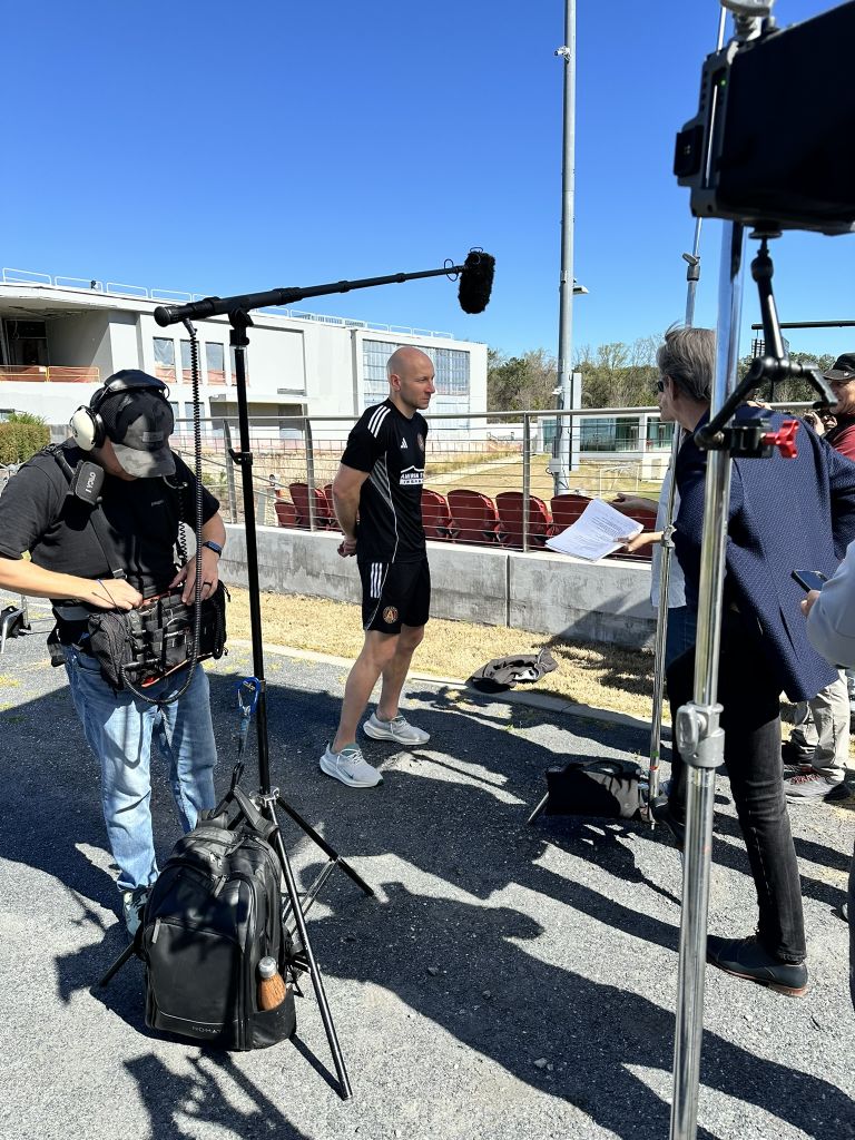A film crew is setting up equipment to interview a man in a sports uniform outdoors in Atlanta, Georgia.