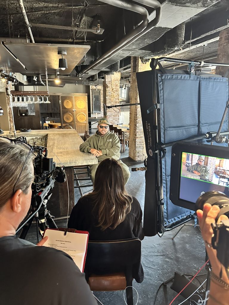 A film crew is setting up to interview a man in a green hoodie in a rustic indoor space in Atlanta, Georgia.
