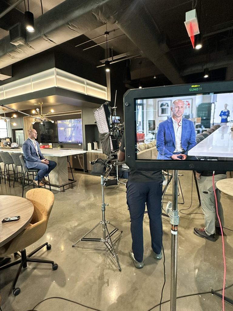 United We Roar 6 A man in a blue suit sits on a barstool while being filmed by a camera crew in a modern office setting in Atlanta, Georgia.