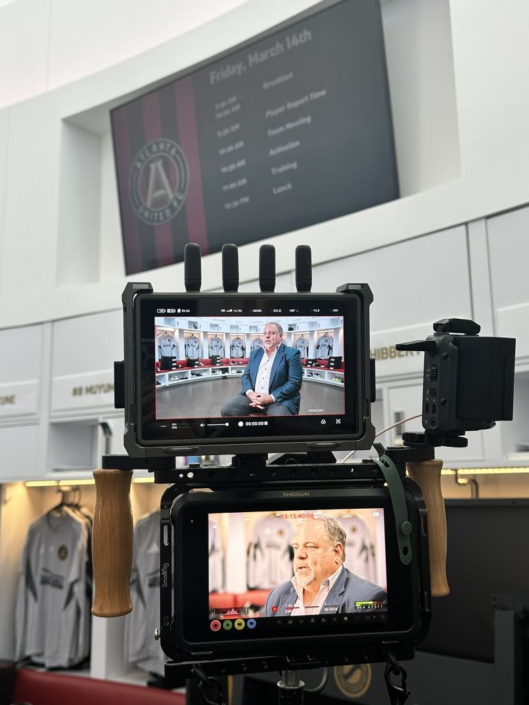Two video cameras recording a man in a suit sitting in a locker room with soccer jerseys in the background in Atlanta, Georgia.
