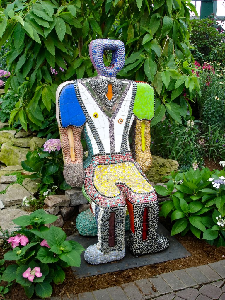 A vibrant, mosaic-covered statue of a humanoid figure sits amidst a lush garden, surrounded by colorful flowers and green foliage in Atlanta, Georgia.
