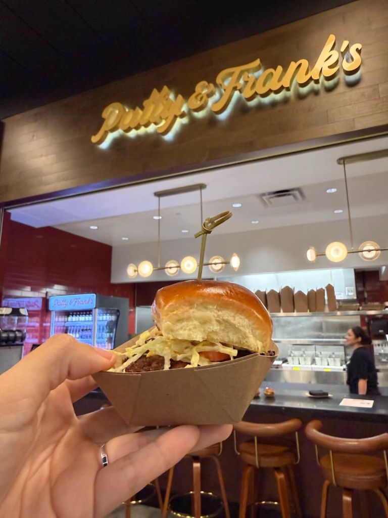 A hand holds a small cardboard tray with a burger topped with lettuce and tomato from Patty & Frank's restaurant in Atlanta, Georgia.