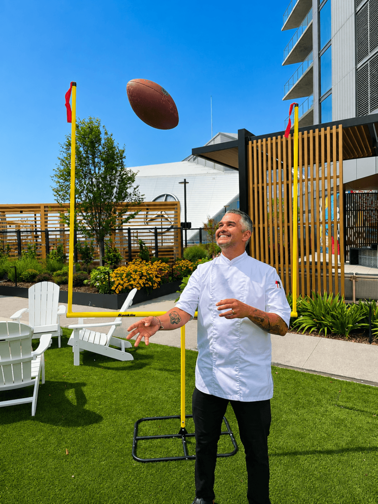 A chef in a white coat throws a football between two yellow goalposts on a grassy rooftop garden in Atlanta, Georgia.