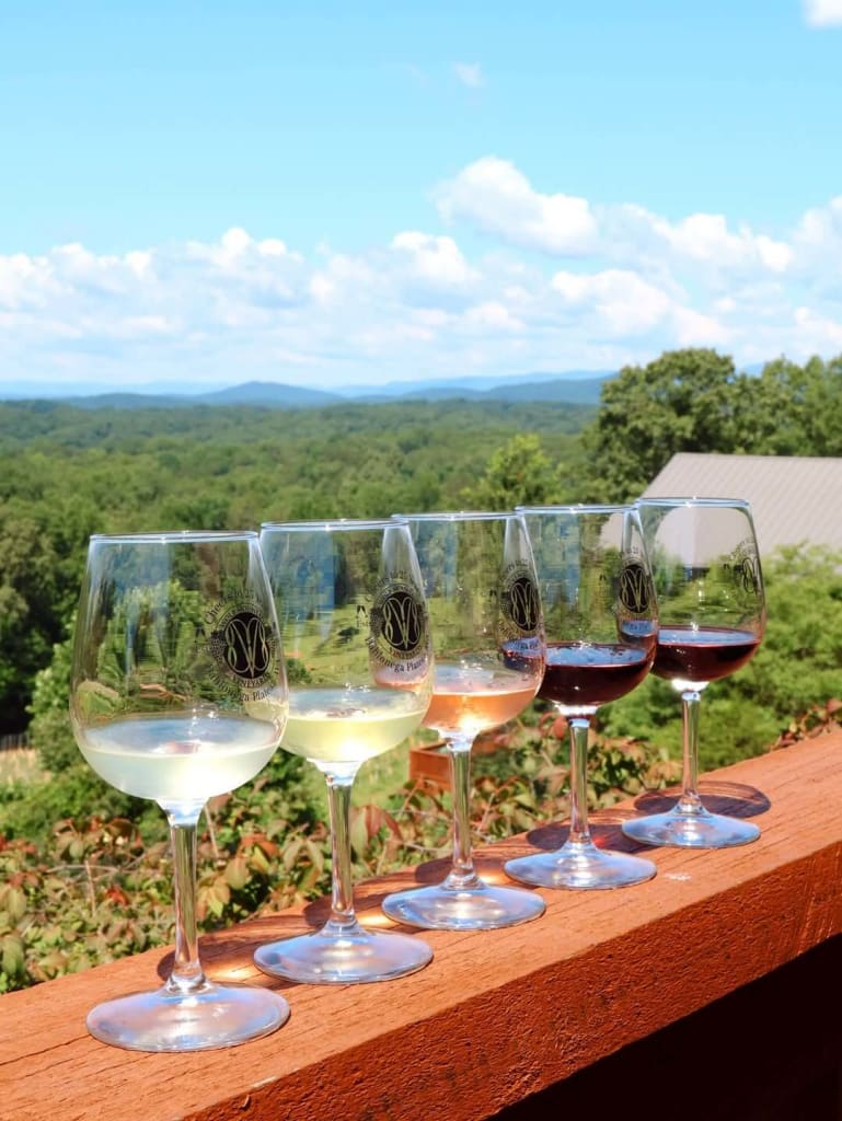 Nearby Day Trips 5 Five wine glasses with different colored wines sit on a wooden railing overlooking a scenic valley in Atlanta, Georgia.