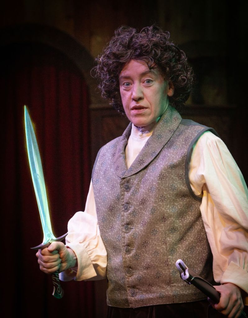 A person dressed in period costume holds a glowing sword and a dagger, standing against a dark, theatrical backdrop in Atlanta, Georgia.