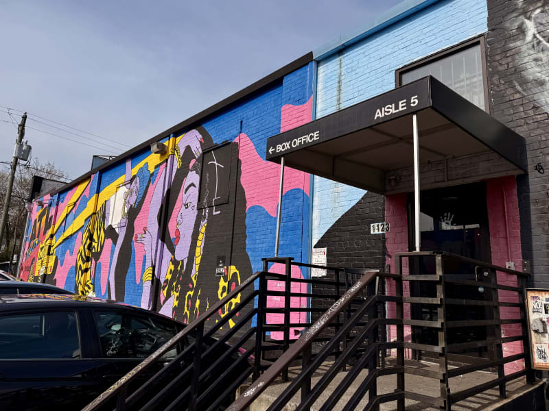 A vibrant mural adorns the side of a building, featuring abstract shapes and figures in bold colors like pink, blue, and yellow, with a sign above a pink door reading 'Aisle 5 Box Office' in Atlanta, Georgia.