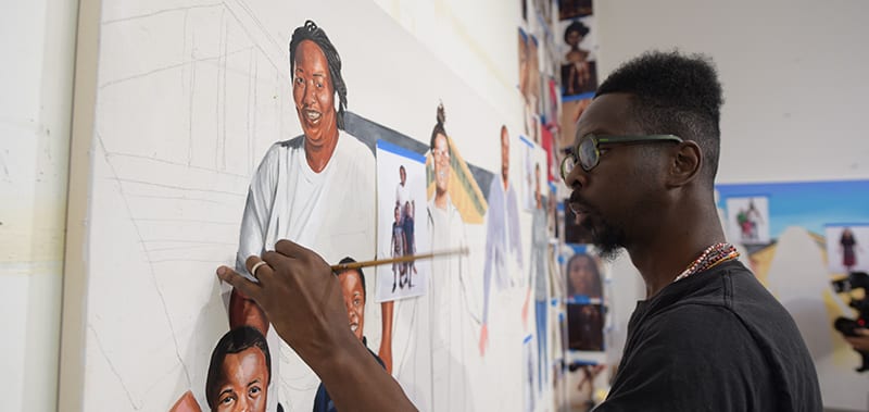 Fahamu Pecou 3 A man paints a mural featuring portraits of smiling people, including children and adults in Atlanta, Georgia.