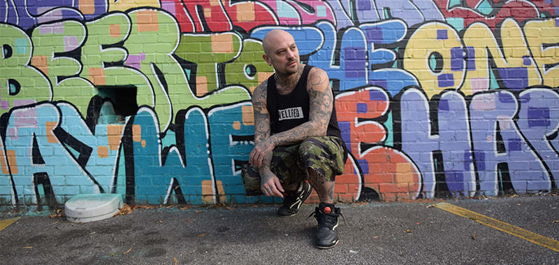 Dr. Dax 3 A bald man with tattoos sits against a colorful graffiti wall in Atlanta, Georgia.