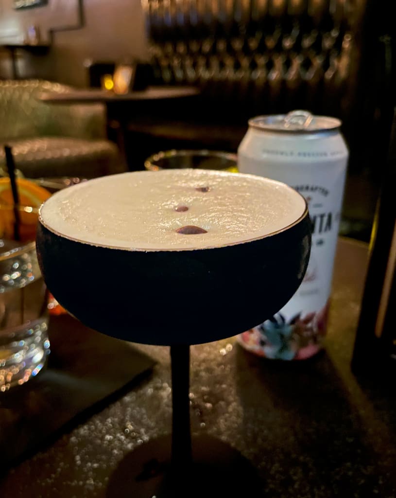 A dark cocktail glass filled with a creamy white drink sits on a table next to a can of beer in Atlanta, Georgia.