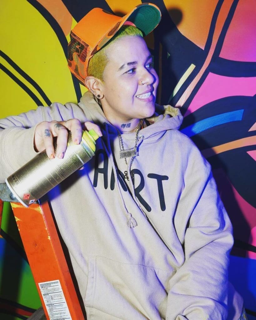 A person with short, bright hair wearing a hoodie and cap sprays paint on a colorful mural in Atlanta, Georgia.