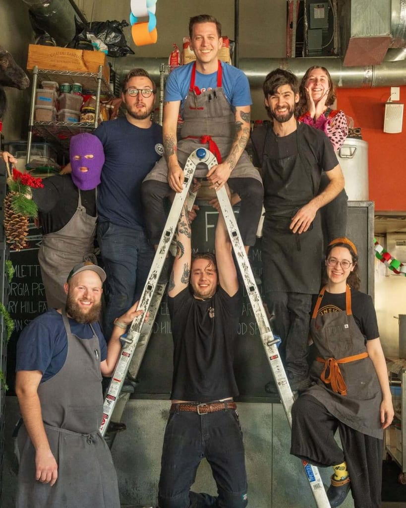 Inside Gunshow Restaurant: How Chef Cody Chassar Leads an Atlanta Icon 3 A group of eight kitchen staff members pose playfully on a ladder in their restaurant kitchen in Atlanta, Georgia.
