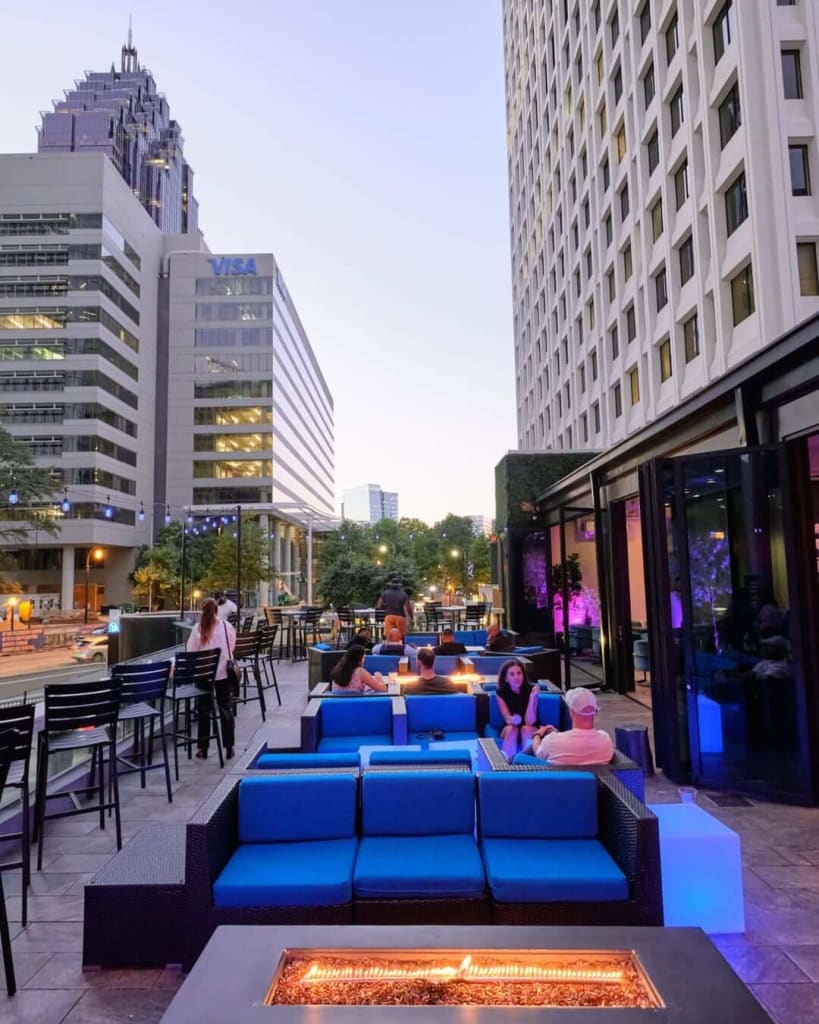 Best Outdoor Patios for Winter Dining 3 People relax on blue couches around a fire pit on a rooftop patio with city skyline views in Atlanta, Georgia.
