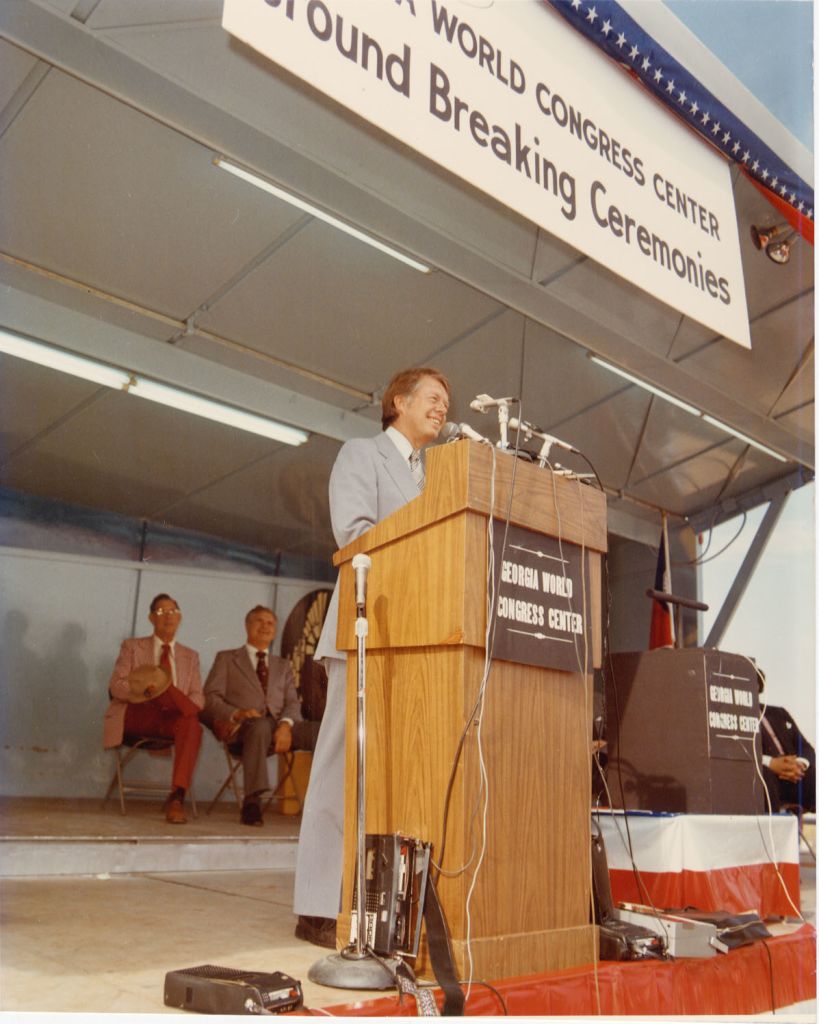 A man in a suit stands at a podium with microphones, addressing an audience at an outdoor event in Atlanta, Georgia.