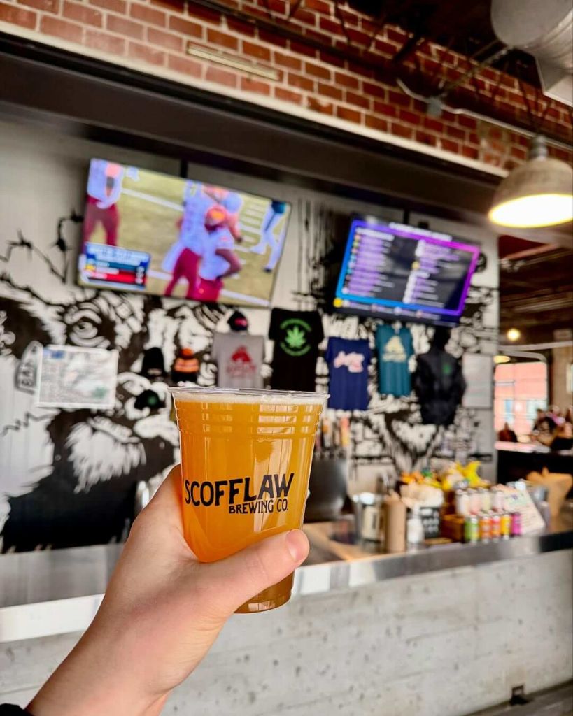 Where to Watch College Football in Atlanta 3 A hand holds a plastic cup of beer from Scofflaw Brewing Co. in a bar with TVs and a brick wall in Atlanta, Georgia.