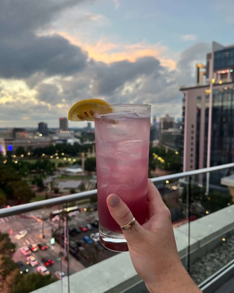 A hand holds a tall glass filled with a vibrant pink cocktail, garnished with a lemon wedge, against a backdrop of a city skyline at dusk with a cloudy sky in Atlanta, Georgia.
