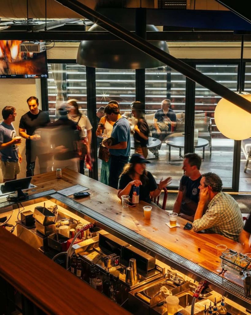 A lively bar scene with patrons enjoying drinks and conversation, illuminated by warm lighting and framed by a large window in Atlanta, Georgia.