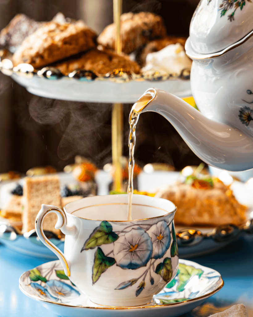 A delicate white teacup adorned with blue and green floral patterns is being filled with steaming tea from a beautifully decorated teapot, set against a backdrop of a lavish afternoon tea spread with scones, pastries, and sandwiches in Atlanta, Georgia.