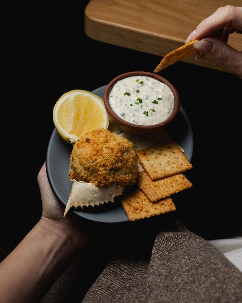 A hand holds a plate with a crispy crab cake, lemon wedge, creamy sauce, and crackers for dipping in Atlanta, Georgia.