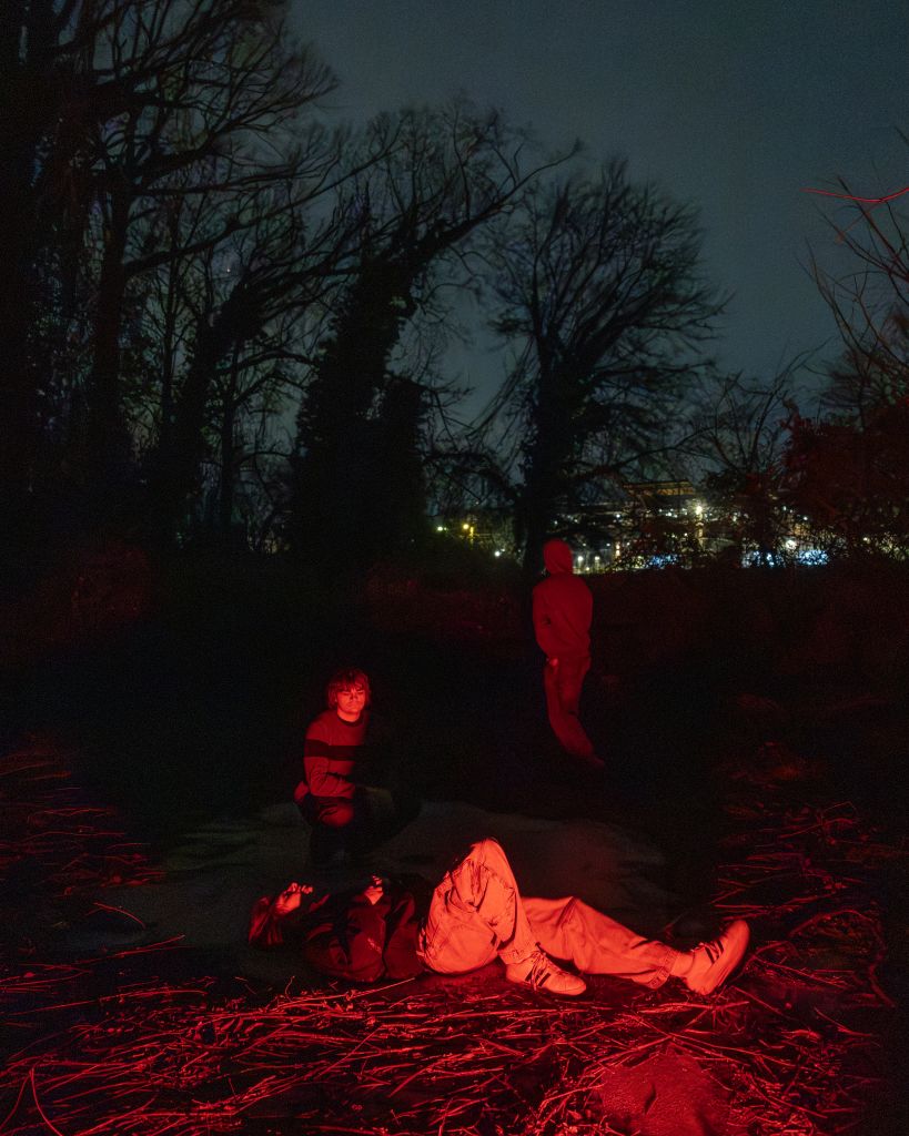 Three individuals are gathered in a dark, wooded area at night, illuminated by a red light that casts eerie shadows and highlights their silhouettes against the backdrop of leafless trees and distant city lights in Atlanta, Georgia.