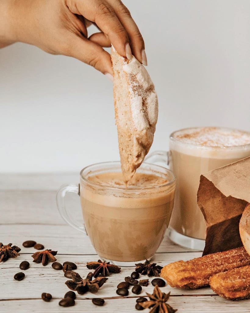 A hand dips a sugar-coated cookie into a frothy latte, surrounded by coffee beans and pastries in Atlanta, Georgia.