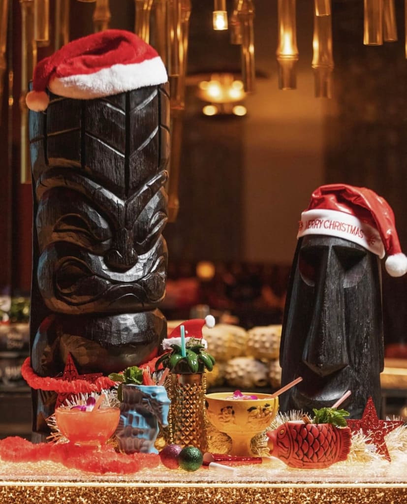 Two large tiki statues, one wearing a Santa hat, stand behind festive holiday cocktails in Atlanta, Georgia.