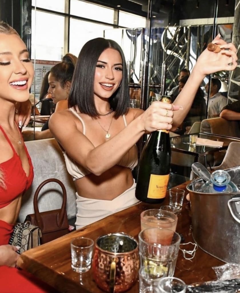 Two women celebrate at a bar, one opening a bottle of champagne while the other smiles in Atlanta, Georgia.