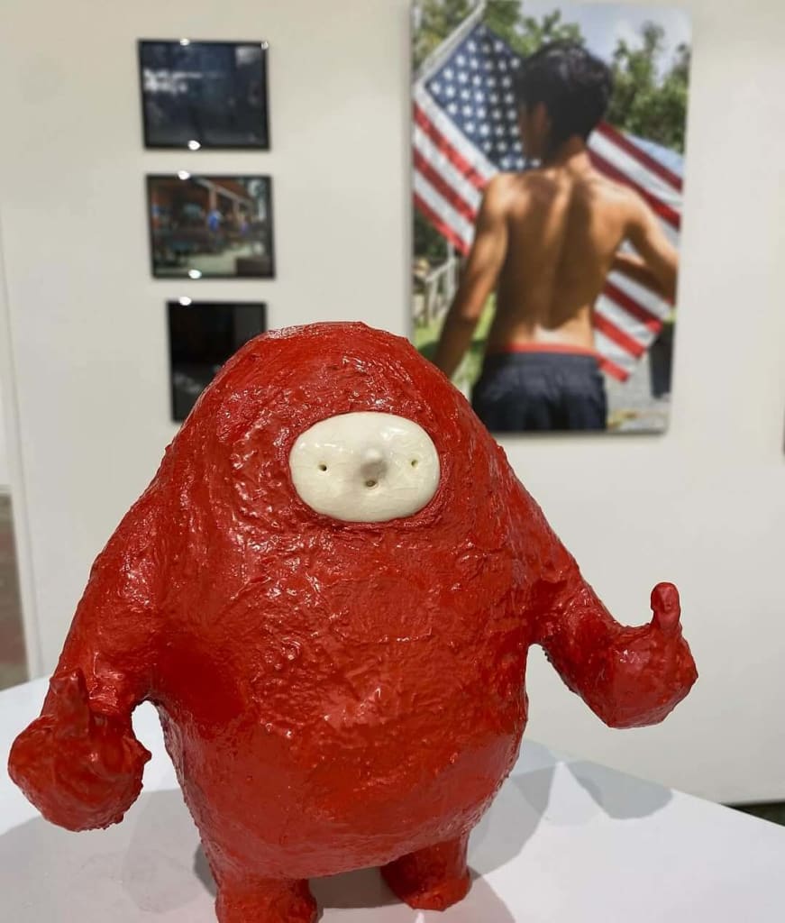 A red, blob-like statue with a white face stands in front of an American flag painting in Atlanta, Georgia.