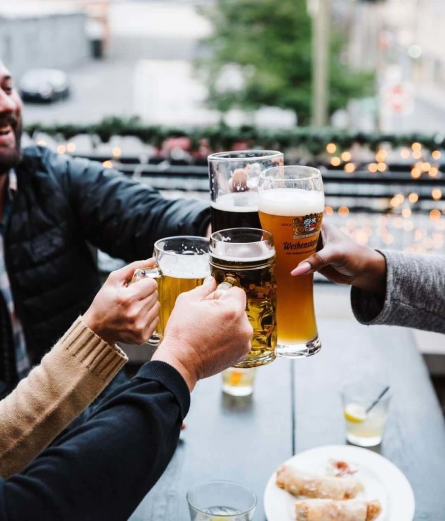 Four friends raise their glasses of beer in a toast on a rooftop patio, surrounded by twinkling lights and a festive atmosphere in Atlanta, Georgia.