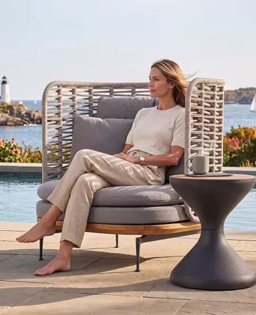 Woman relaxing in a woven lounge chair beside a pool with a coastal view.