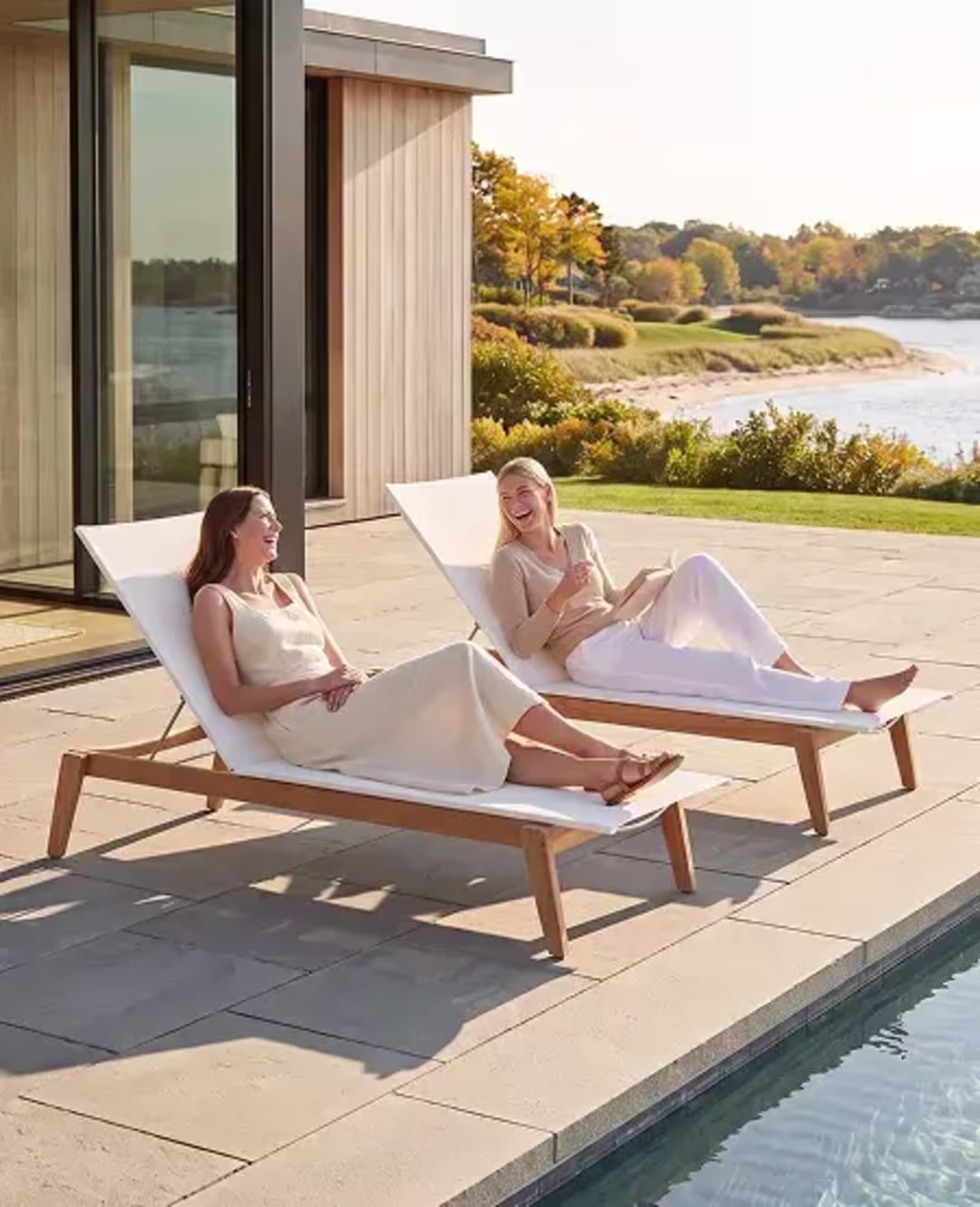 Two women relaxing on chaise lounges beside a modern pool overlooking the coast.