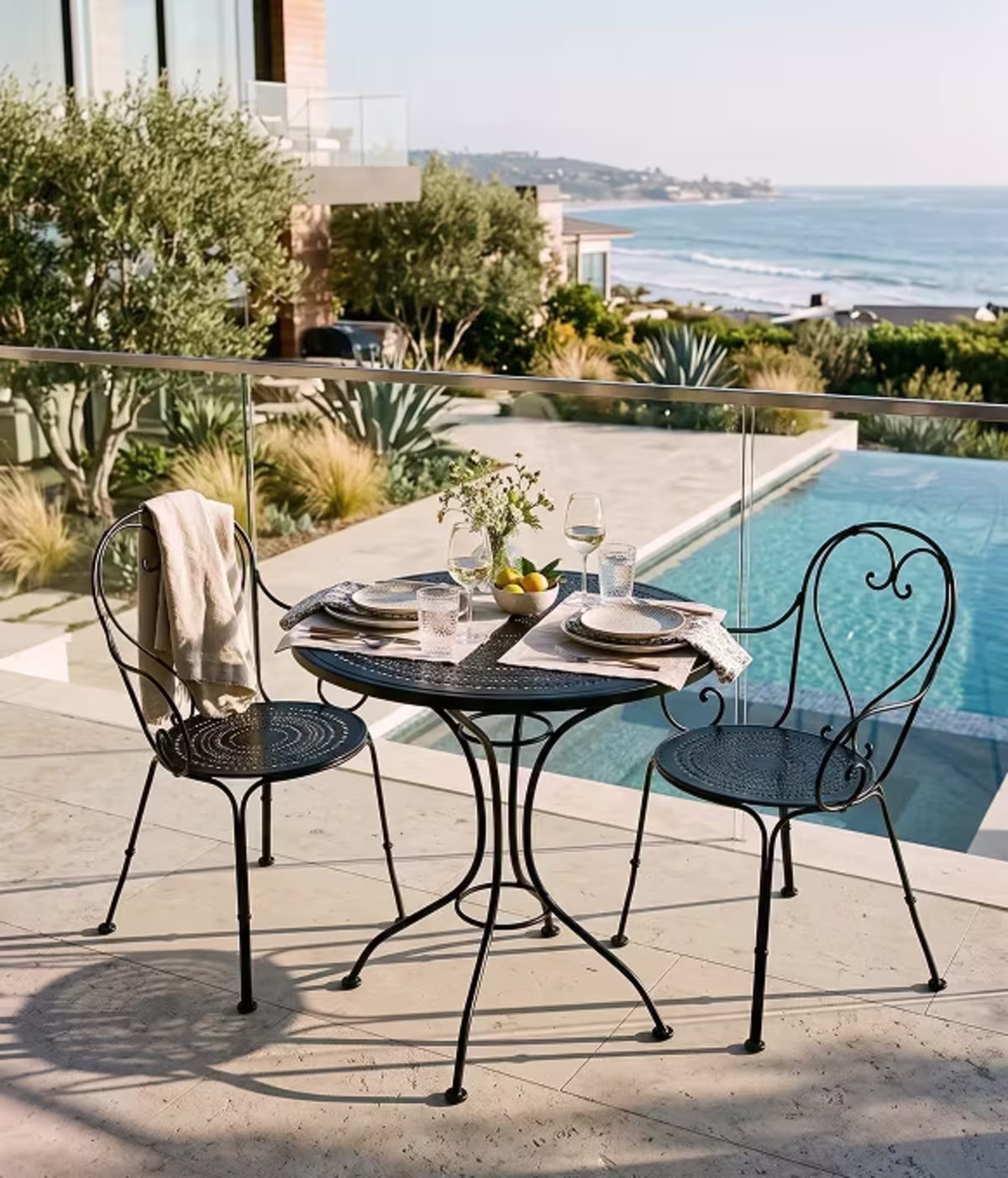 Image of a black wrought iron bistro dining set with elegant place settings beside a pool overlooking the ocean.