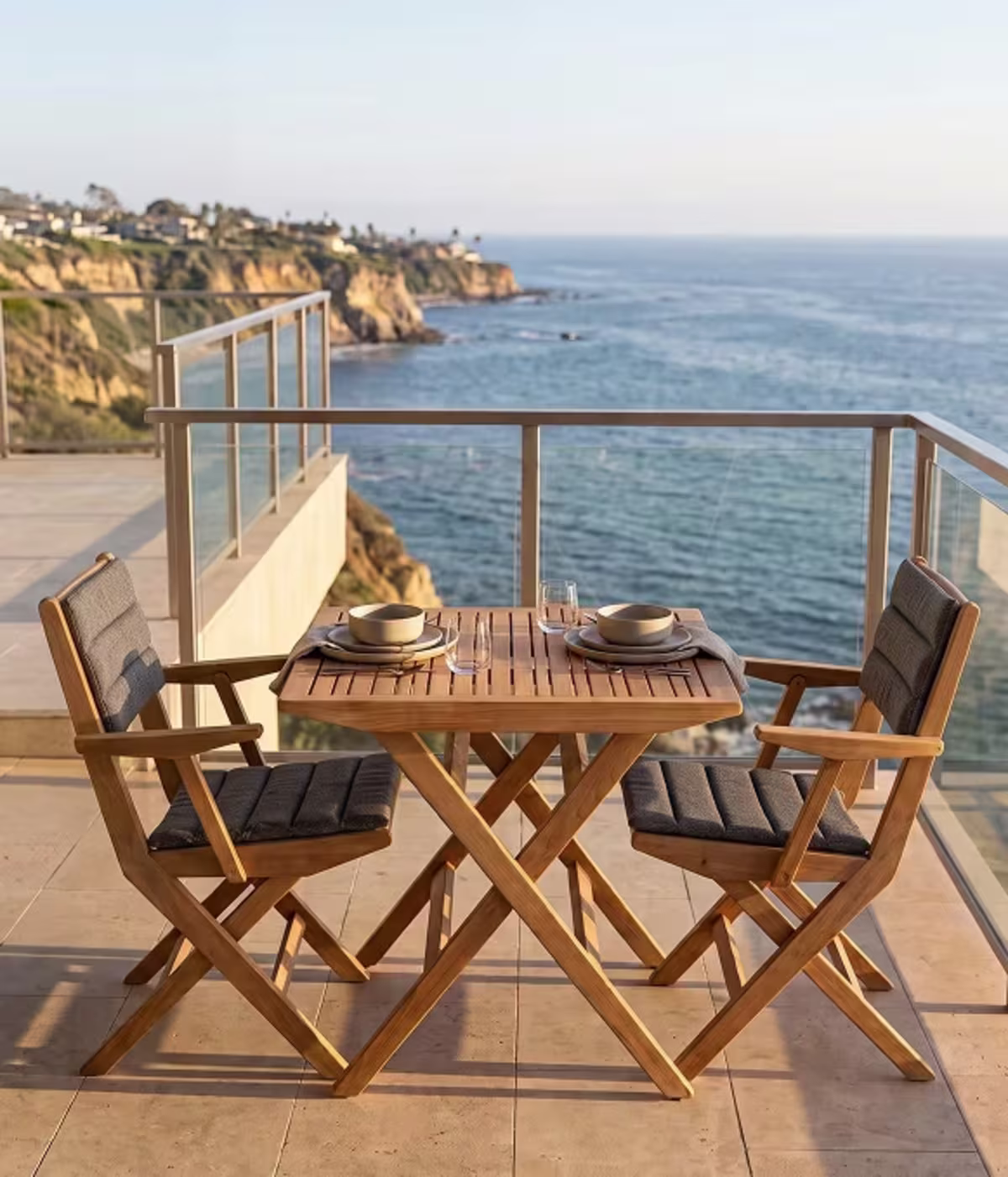 Image of a folding teak bistro dining set with dark gray cushions and two place settings on an oceanfront balcony.