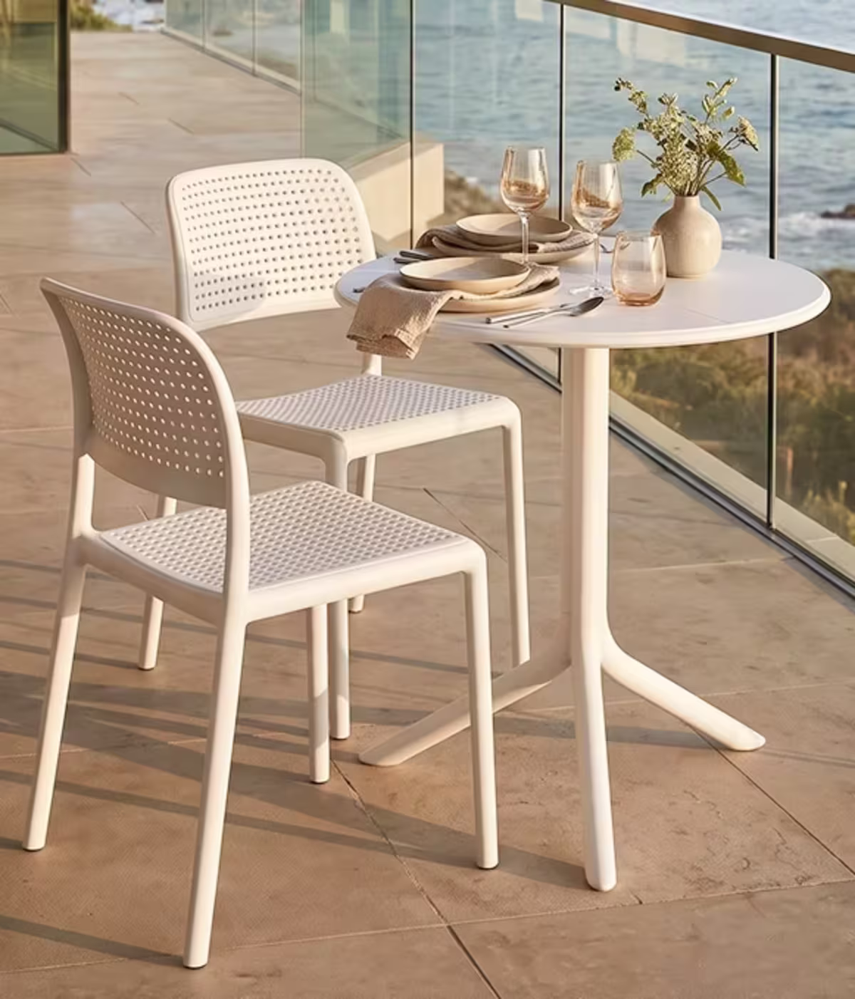 Image of a white round bistro table with two perforated chairs, elegant place settings, and flowers overlooking the ocean.