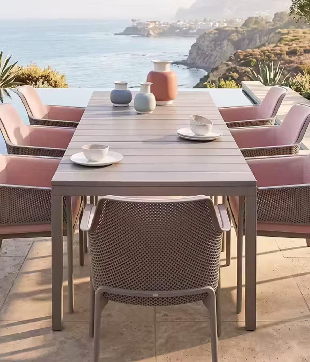 Modern rectangular outdoor dining table with woven chairs and pastel accents overlooking the coast.