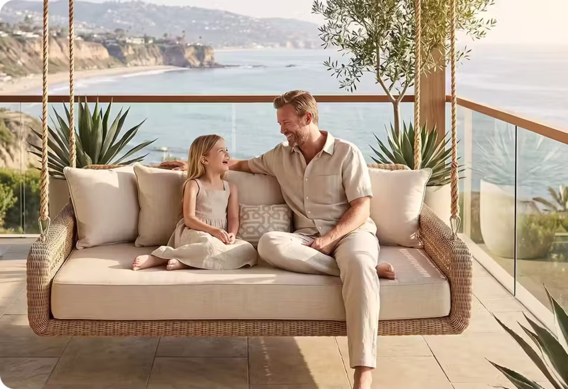 Father and young daughter smiling together on a woven outdoor hanging daybed overlooking the ocean from a contemporary patio. image